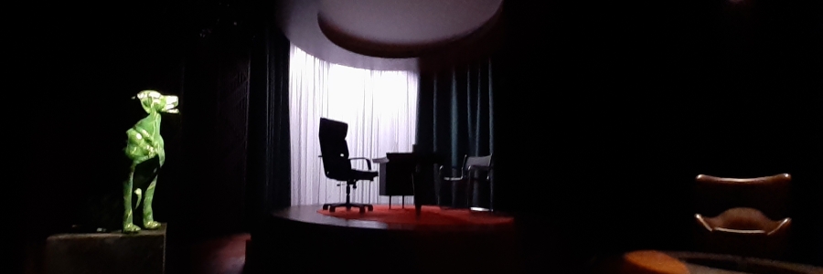 Hämärä teatterin näyttämö. Vasemmalla valokeilassa vihreä koirapatsas. Keskellä korokkeella konttoripöytä ja kaksi tuolia. Oikeassa laidassa ruskea nahkatuoli.