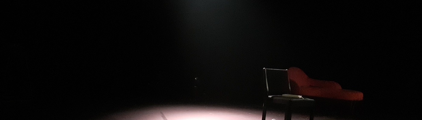 Musta lava, jonka keskellä valo valaisee mustan tuolin, jonka alla on juomapullo. Taustalla näkyy punainen divaani.